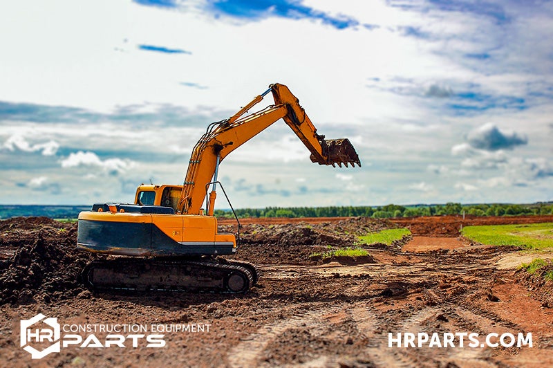 An excavator works in a field with trees in the distance and under a warm sky with soft, fluffy clouds.